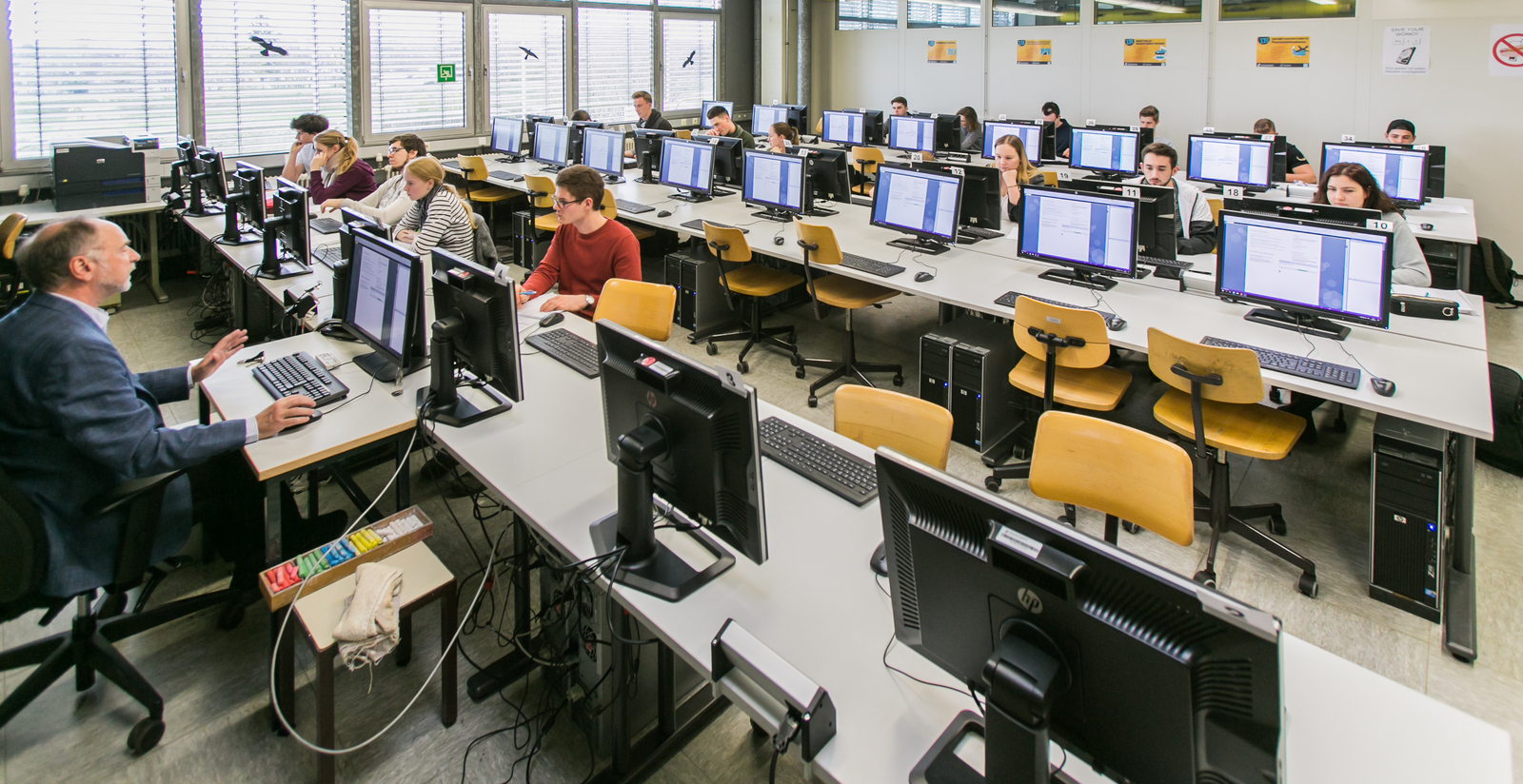 Ein Professor und Studierende sitzen in einem Hörsaal an Computern