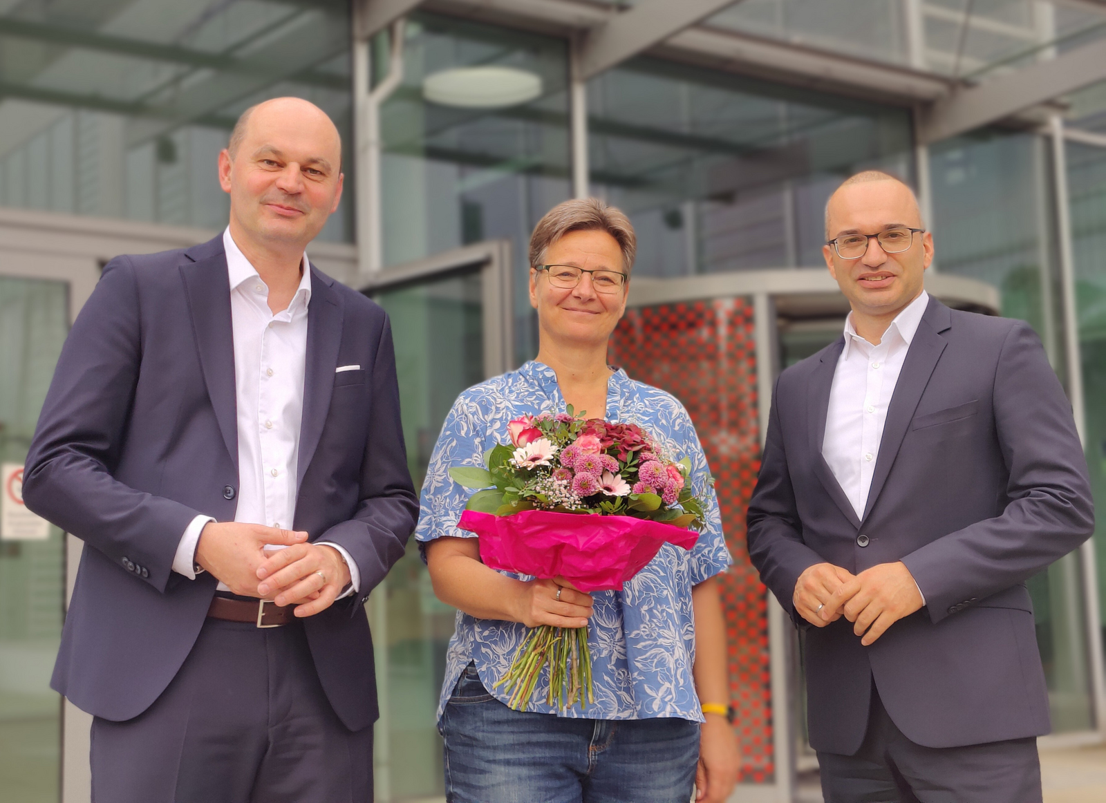 Die drei Personen stehn vor Gebäude D, Grit Köhler hält einen Blumenstrauß in der Hand