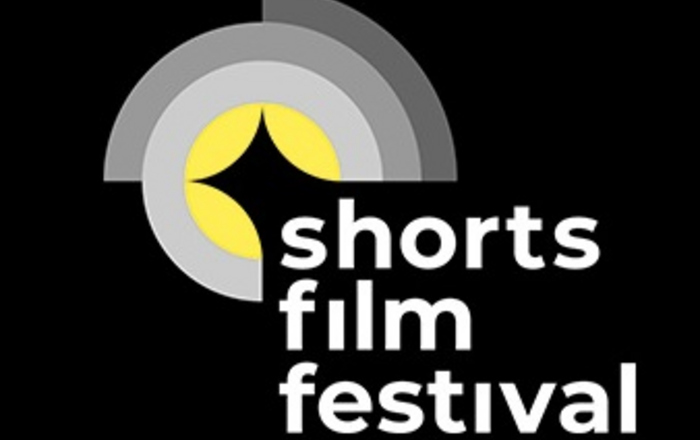 Logo des shorts film festivals in den Fraben scharz weiß grau und gelb deutet durch mehre ineinander liegende Halbkreise und Kreise ein Kameralinse an in deren unterer  rechter Ecke untereinander die Wort shorts film festival stehen 