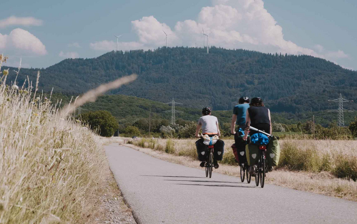 Filmteam auf radelt auf Landstraße durch Kornfelder im Hintergrund ein Schwarzwaldberg.