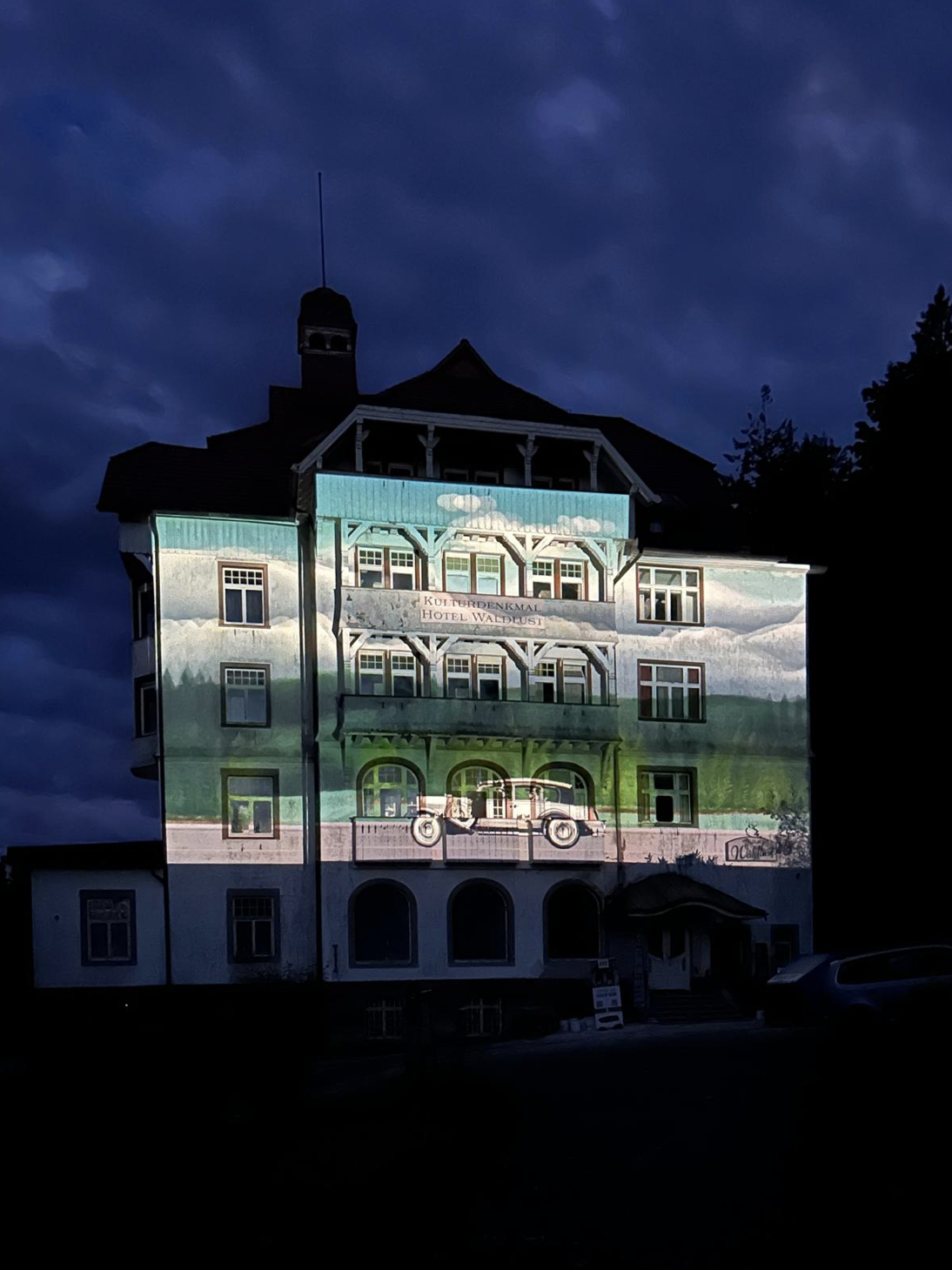 Das ehemalige Grandhotel Waldlust bei Nacht an die Außenfassade ist eine LAndschaft und darin ein Oldtimer-Auto projiziert